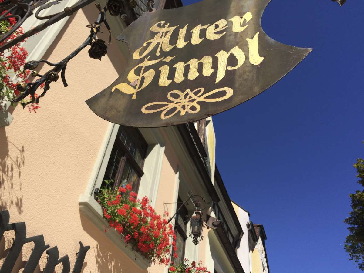 Das traditionelle Wirtshausschild des Restaurants "Alter Simpl" hängt an einer Fassade mit roten Blumen.
