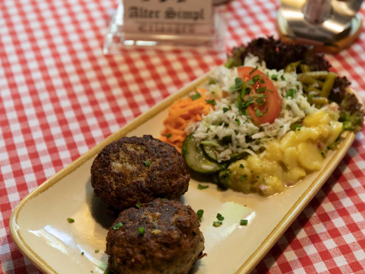 Zwei hausgemachte Fleischküchle auf einem länglichen Teller, dazu gemischter Salat, Kartoffelsalat und eine Tomatenspalte.