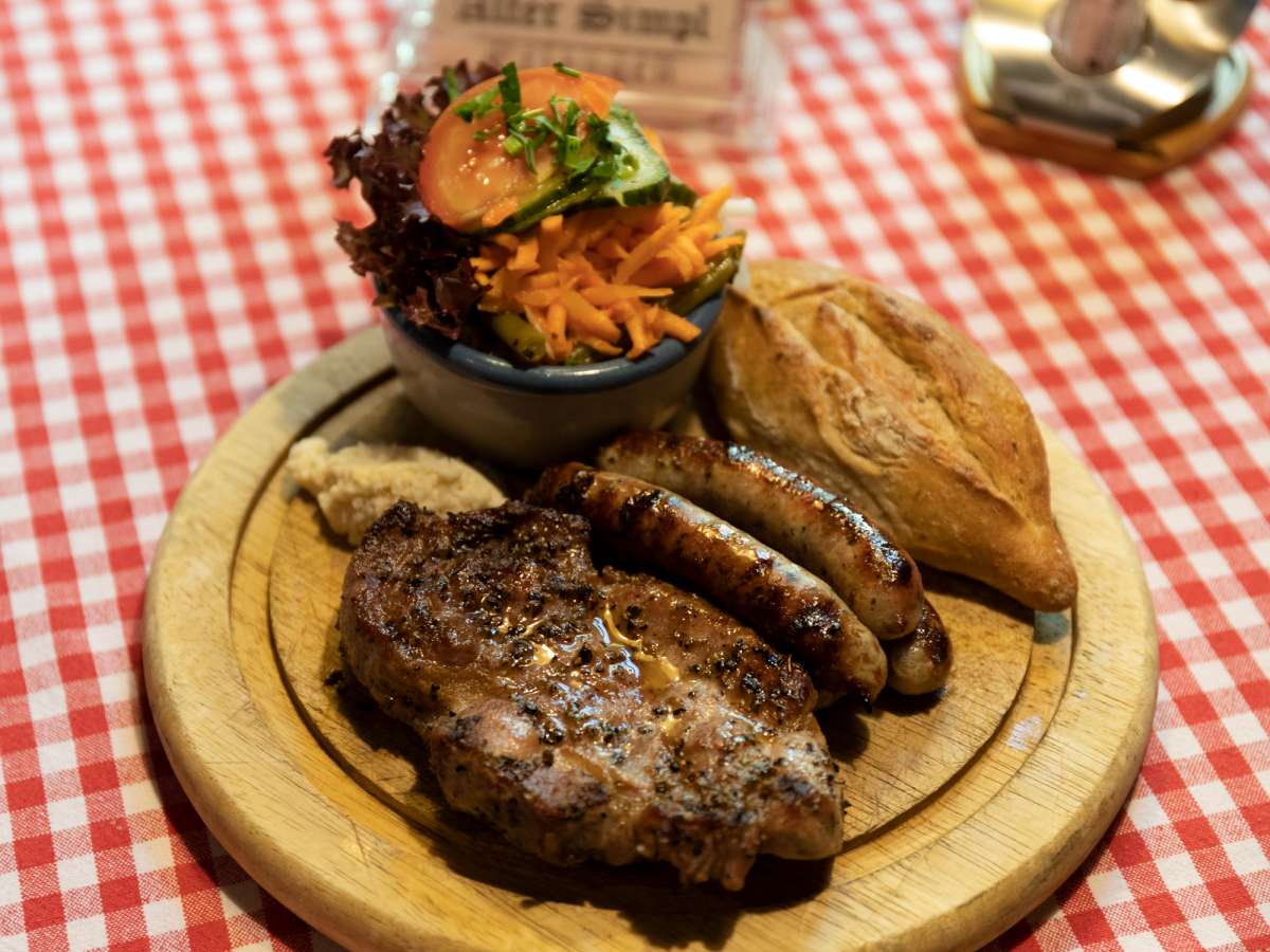 Der Mixer auf dem Holzbrett: eine Scheibe Grillfleisch vom Buchenholzgrill mit Original-Rostbratwürsten, einer Schale gemischtem Salat und einem Roggenbrötchen.