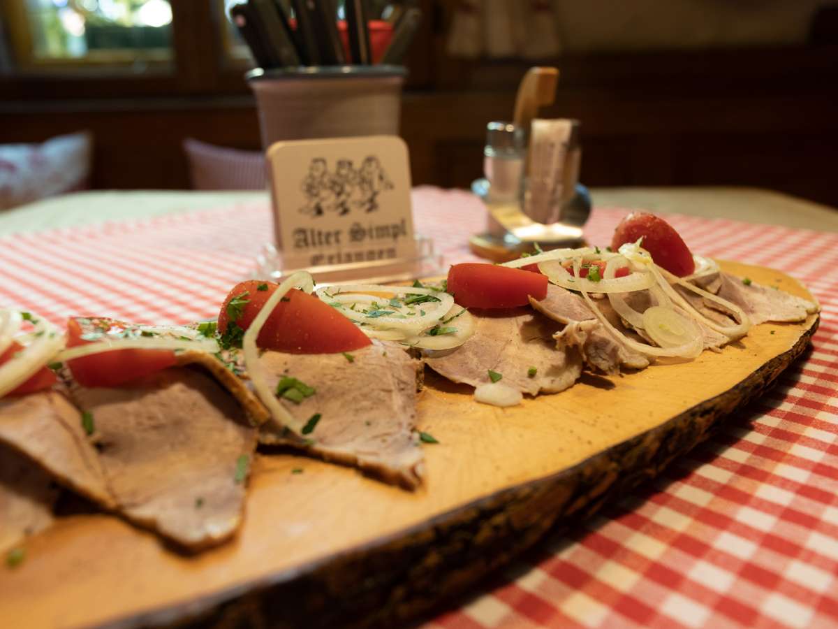 Das Spezialbrot „Alter Simpl“ auf dem Holzbrett: Bauernbrot mit Obatzter, dünn aufgeschnittenem kaltem Braten, Tomate, Zwiebel und Petersilie.