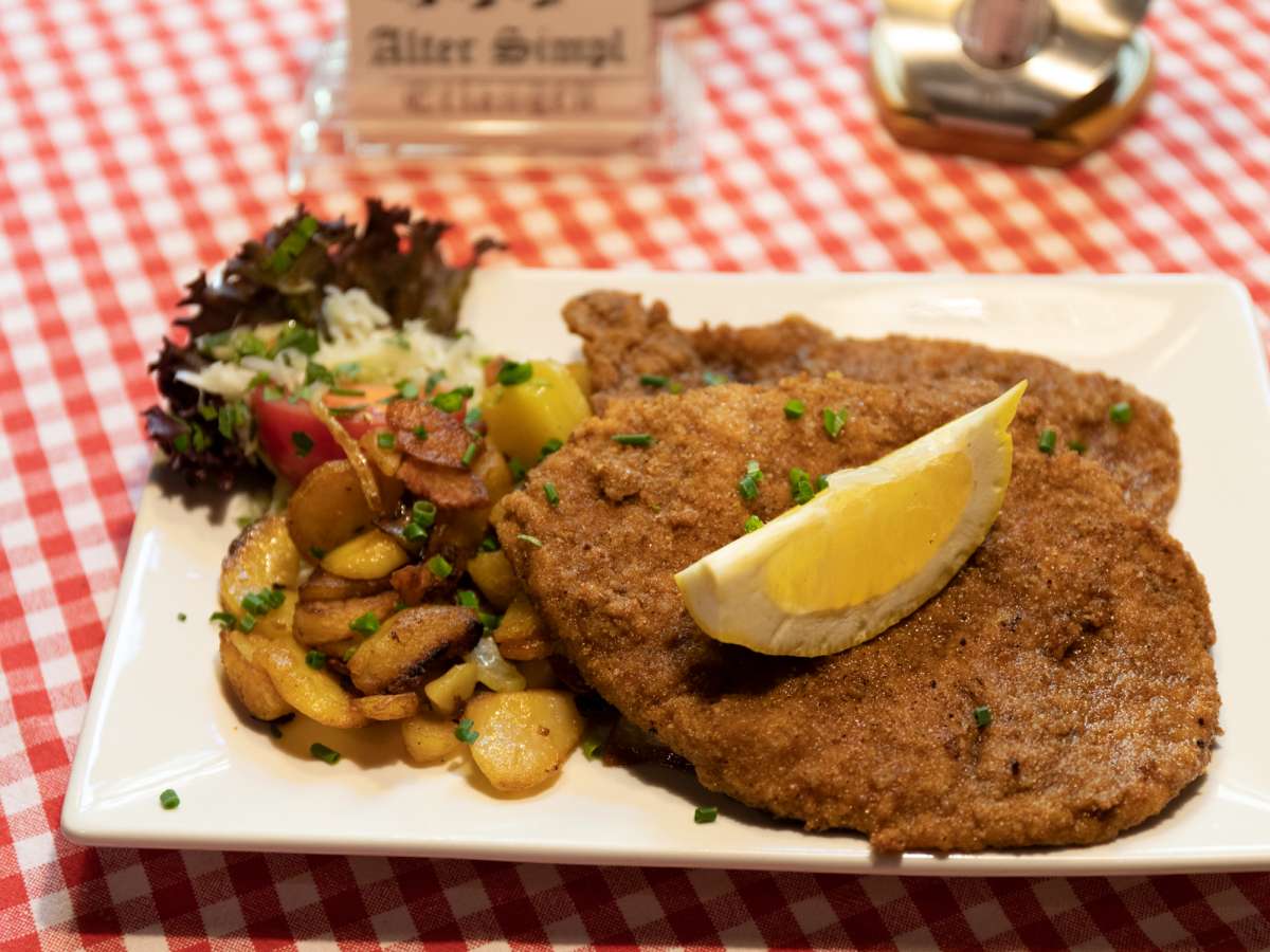 Paniertes Schnitzel Wiener Art aus der Pfanne mit Bratkartoffeln, Zitronenspalte und einem kleinen Salatbukett.