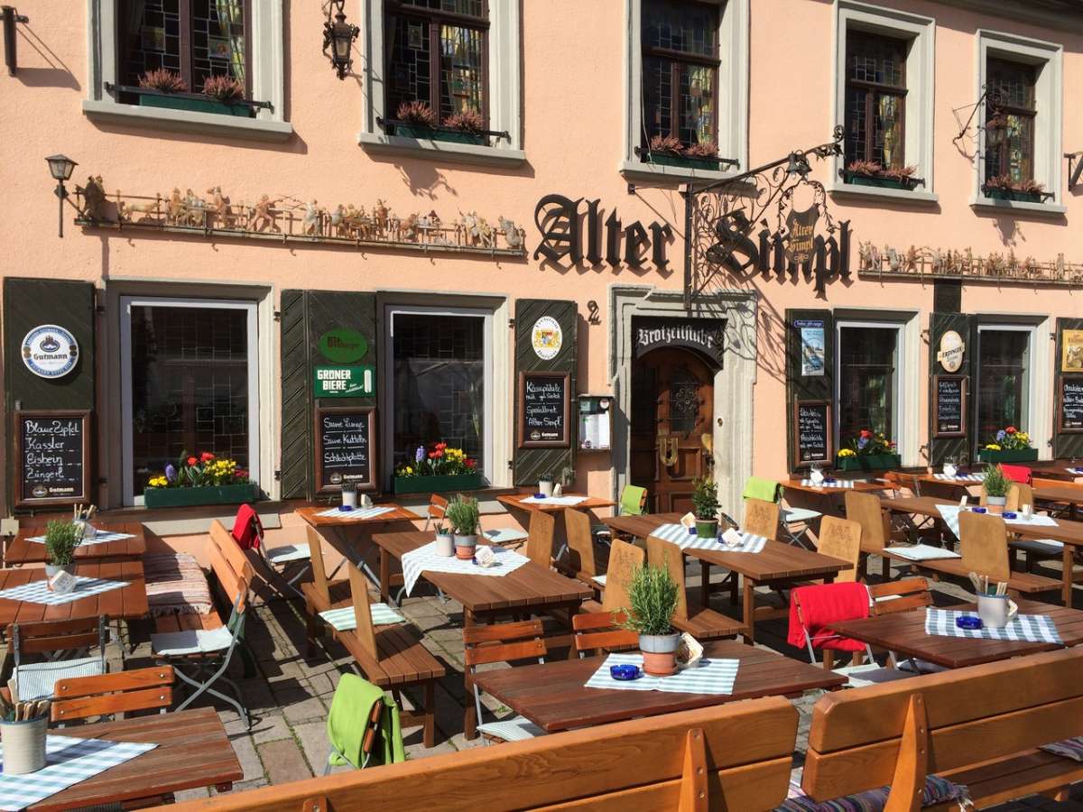 Der Biergarten vor dem Alten Simpl an einem sonnigen Tag: eingedeckte Holztische vor der lachsfarbenen Fassade, darüber das schmiedeeiserne Wirtshausschild und geschnitzte Figuren, an den Fensterläden Tafeln mit Tagesgerichten.