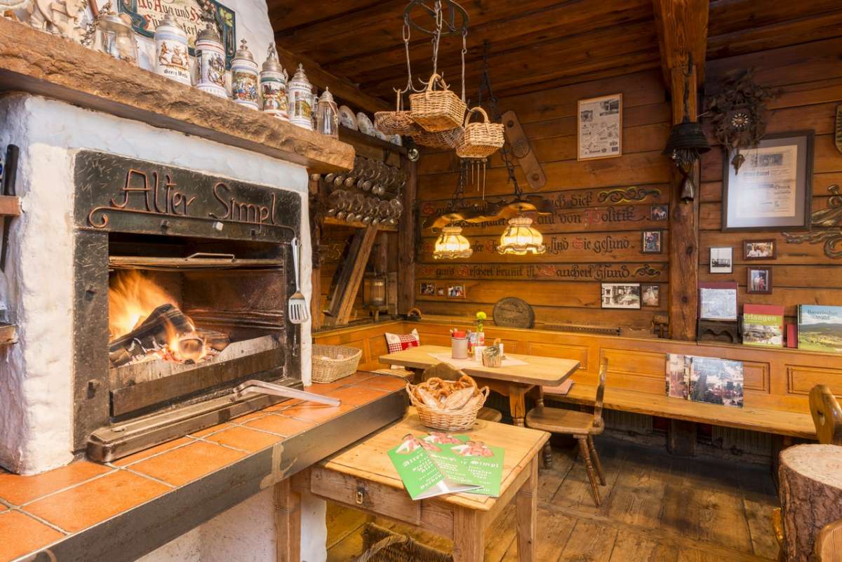 Der Buchenholzgrill mit offenem Feuer, in die Haube ist „Alter Simpl“ geschmiedet; darüber eine Reihe Bierkrüge, daneben ein Korb mit Brezen und Bauernbrot in der holzvertäfelten Stube.
